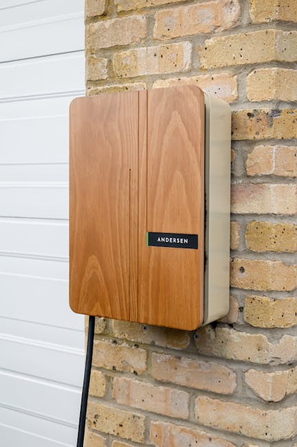 A close-up view of a wooden outdoor electrical cabinet mounted on a brick wall near a white garage door, with a black power cable connected to it. The cabinet has a smooth, light wood finish with a black label reading 'ANDERSEN' attached to its front. The setting appears to be part of a home exterior, possibly during an initial stage of loading or staging for house removals by Man With a Van Crayford. The image captures a well-maintained brick wall contrasting with the sleek, modern appearance of the cabinet, which is positioned at eye level. The surrounding environment suggests preparation for moving or packing activities, aligned with furniture transport and home relocation services. The natural daylight and neutral tones emphasize the clarity of the objects involved in the loading process within a residential setting, illustrating part of the logistical steps in professional removals.