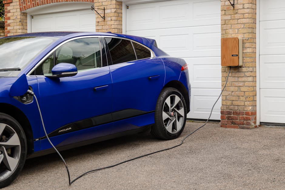 Close-up view of an electric vehicle charging station situated outdoors next to green leafy bushes, with multiple charging cables plugged into the station's ports. The station has a blue and white design, featuring labels and icons indicating it is an electric vehicle charger, including a prominent charging symbol. Two white bollards with red stripes are positioned in front of the station to protect it from vehicle collision. The setting is well-lit by natural daylight, illustrating the charging infrastructure associated with home relocation or moving logistics services offered by Man With a Van Crayford, supporting environmentally conscious furniture transport and packing and moving procedures.