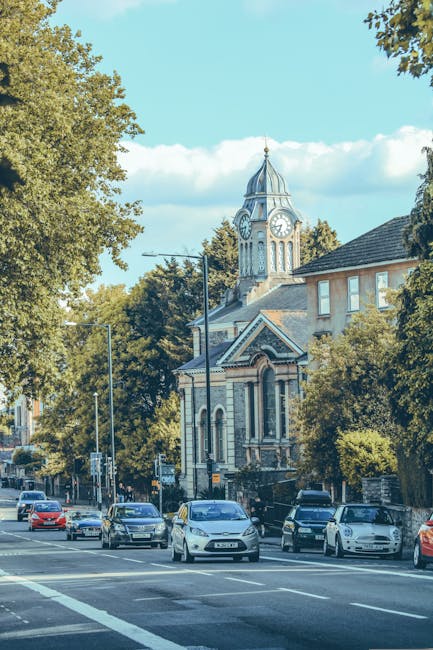 A row of parked cars along a city street with a church featuring a tall clock tower and a spire visible in the background. The street is lined with large leafy trees providing shade, and there are lampposts along the pavement. The area appears calm with no pedestrians or moving vehicles in sight. The scene is captured during daylight with clear blue skies and some clouds. This setting illustrates a typical urban environment where house removals and furniture transport services by Man With a Van Crayford may operate, especially when adhering to local council permit rules for street parking and loading during home relocation process. The image emphasizes the importance of planning for vehicle access and parking during house moves in the area.