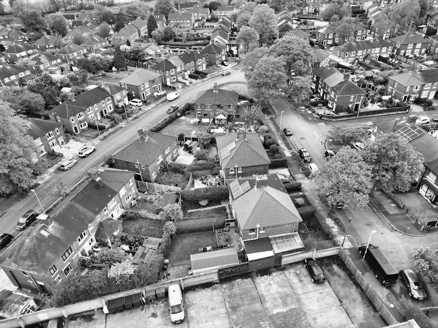 An aerial view of a residential neighbourhood in Crayford, showing a mixture of detached and semi-detached houses with pitched roofs, gardens, and parked cars along the streets. Several houses feature small backyard gardens with fencing, some containing outdoor storage sheds, patio furniture, and garden plants. The image captures a moment during a home relocation process where a manwithavancrayford.co.uk crew is likely involved in furniture transport or packing and moving activities, evidenced by the presence of a large black van parked on the street, adjacent to a driveway with items possibly prepared for loading. Surrounding roads are lined with trees, providing partial canopy over the area. There is clear space on the pavement for loading or unloading, and the environment is well-lit with natural daylight. This scene highlights typical moving logistics in a suburban setting, emphasizing the planning and coordination required for house removals in the area.