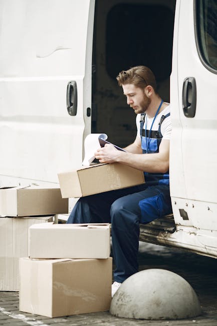 A man wearing a white t-shirt, jeans, and a black cap loading several cardboard boxes into the side of a white delivery van parked on a residential street. The van has its rear doors open, with additi