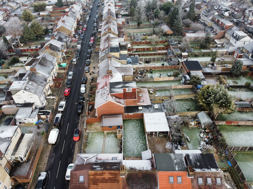 An aerial view of a residential street in Crayford, showing terraced houses lined along a narrow asphalt road with parked cars on either side. The houses are primarily brick-built with pitched roofs, some having snow or frost on the rooftops indicating winter weather. Behind each property, there are fenced back gardens, mostly enclosed with wooden or brick fences, featuring lawn areas, garden sheds, and outdoor furniture. In some backyards, visible cardboard boxes, packing materials, and household items suggest packing and home relocation preparations. The street scene captures the typical environment for house removals, with close proximity between properties and access to rear gardens, which can pose challenges in furniture transport and loading during end terrace removals. Occasionally, workmen may be seen using trolleys or moving equipment to facilitate the loading process, aligning with services like those provided by Man With a Van Crayford, specializing in removals and local home moving logistics.