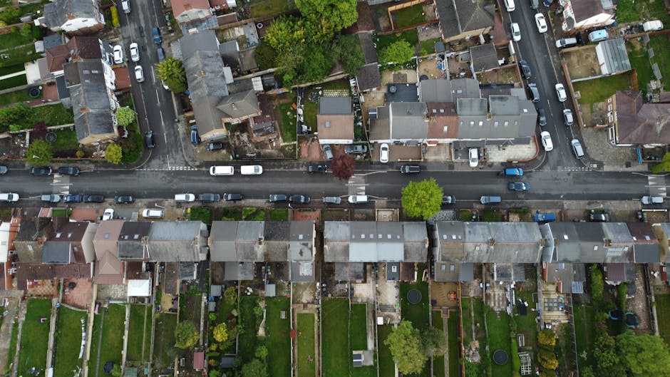 An aerial view of a residential street in Crayford, showing terraced houses lined along a narrow asphalt road with parked cars on either side. The houses are primarily brick-built with pitched roofs, some having snow or frost on the rooftops indicating winter weather. Behind each property, there are fenced back gardens, mostly enclosed with wooden or brick fences, featuring lawn areas, garden sheds, and outdoor furniture. In some backyards, visible cardboard boxes, packing materials, and household items suggest packing and home relocation preparations. The street scene captures the typical environment for house removals, with close proximity between properties and access to rear gardens, which can pose challenges in furniture transport and loading during end terrace removals. Occasionally, workmen may be seen using trolleys or moving equipment to facilitate the loading process, aligning with services like those provided by Man With a Van Crayford, specializing in removals and local home moving logistics.