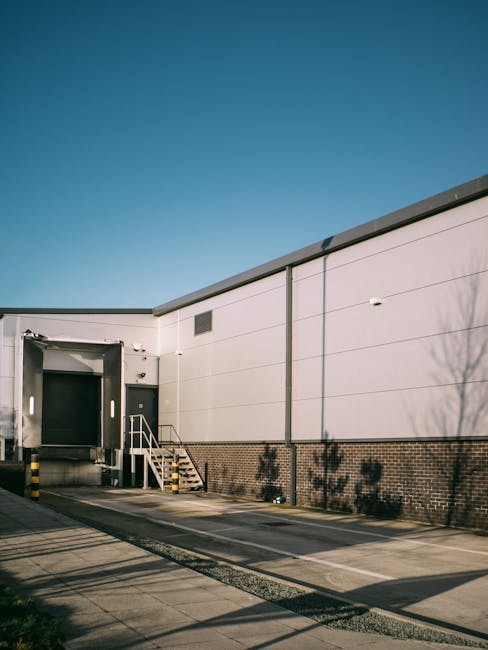 The image shows the exterior of a warehouse or industrial building located on Crayford Industrial Estate, with a clear blue sky overhead. The building's façade is composed of grey metal panels and brickwork at the lower section. To the left, there is a large, dark green loading dock door with a small set of metal stairs and handrails leading up to it, indicating a point for furniture and equipment loading and unloading during a home or office relocation. In front of the building, there is a paved loading area with designated parking spaces, some of which are temporarily blocked by yellow and black safety barriers. The area appears clean and organized, with minimal shadowing from nearby trees, which are also faintly visible on the right side of the image. This setting reflects a typical commercial environment suitable for cargo transport and furniture logistics, and it is associated with professional removals services such as those provided by Man With a Van Crayford, particularly during packing and loading procedures for business or residential moves.