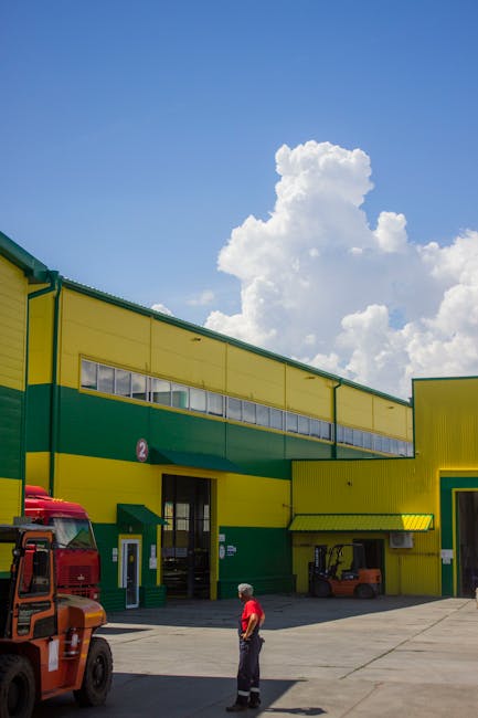 A man in a red shirt and dark trousers stands outside an industrial warehouse on Crayford Industrial Estate during daylight hours. The building features a bright yellow and green exterior with large, rectangular windows near the roofline, and a black doorway beneath a green canopy. To the left, a red truck is partially visible, and a small forklift is parked near the entrance. The sky above is blue with large, fluffy white clouds, indicating fair weather. The scene captures the loading and unloading process typical of commercial removals, with the area clear for vehicle access and movement of furniture or packaging materials. As part of the moving logistics process, [COMPANY_NAME], offered by manwithavancrayford.co.uk, appears to be engaged in loading or preparing items for transportation, reflecting the practical aspects of home or business relocations involving wrapping, packing, and furniture transport on a busy commercial estate.