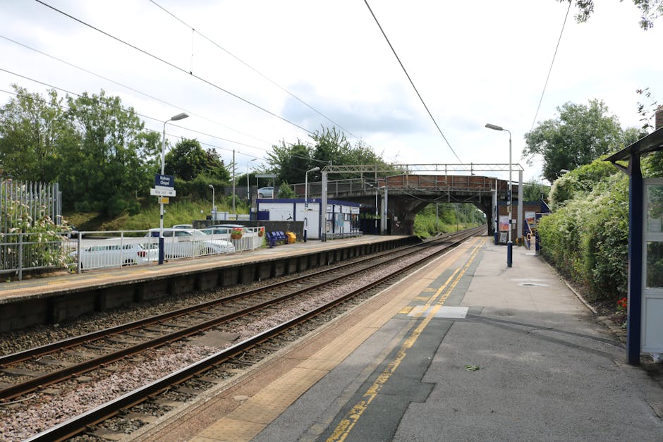 A railway station platform at Crayford with a view towards the train tracks, which run parallel to the paved walkway. On the platform, there are several cardboard boxes of varying sizes, some wrapped in plastic or fabric, likely containing household items for a home relocation. A metal fence separates the platform from a small parking area with white cars visible, and nearby, a blue and white station building can be seen with parking meters and a bus stop shelter on the right side. Overhead, electrical wires stretch across the sky, and a bridge crosses above the tracks, supported by metal frames. The area is outdoors, surrounded by greenery including bushes and trees, with a partly cloudy sky overhead. The scene depicts a typical moving logistics moment where boxes and packing materials are prepared for transport, possibly being loaded onto a vehicle parked just out of view for furniture transport or house removals, with the presence of a company like Man With a Van Crayford implied in the context of this house removals guide.