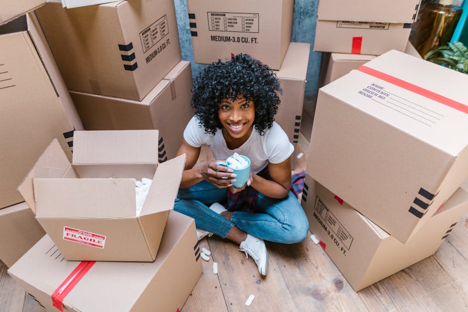 A young woman with curly black hair and a bright smile is sitting cross-legged on a wooden floor inside a property surrounded by numerous cardboard boxes of various sizes, some marked with labels and tape indicating fragile contents and free space for packing. She holds a blue cup with white packing foam inside, smiling at the camera. The boxes are stacked around her, with open boxes revealing packing materials such as tissue paper and foam. Some boxes are positioned on the floor, while others are stacked against the wall, indicating an active packing process in preparation for home relocation. The scene is well-lit with natural light, and the environment suggests a typical moving day with packing materials and furniture ready for transport. This setup illustrates the packing and loading stage of a house removal carried out by specialist moving services such as Man With a Van Crayford, ensuring a smooth furniture transport and efficient home relocation.