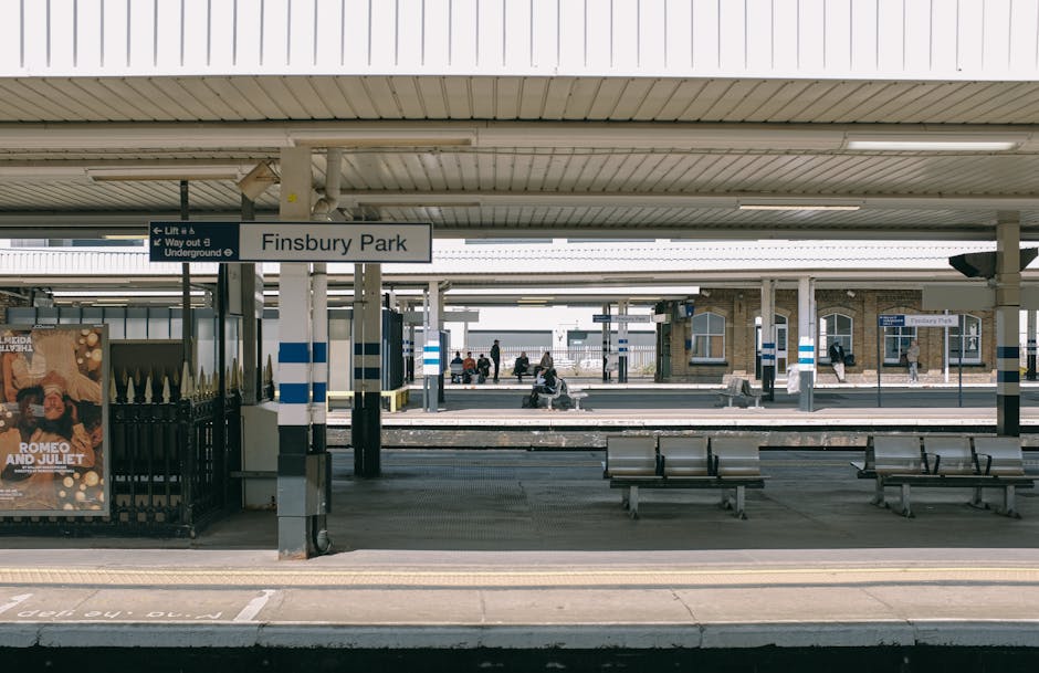 A railway station platform at Crayford with a view towards the train tracks, which run parallel to the paved walkway. On the platform, there are several cardboard boxes of varying sizes, some wrapped in plastic or fabric, likely containing household items for a home relocation. A metal fence separates the platform from a small parking area with white cars visible, and nearby, a blue and white station building can be seen with parking meters and a bus stop shelter on the right side. Overhead, electrical wires stretch across the sky, and a bridge crosses above the tracks, supported by metal frames. The area is outdoors, surrounded by greenery including bushes and trees, with a partly cloudy sky overhead. The scene depicts a typical moving logistics moment where boxes and packing materials are prepared for transport, possibly being loaded onto a vehicle parked just out of view for furniture transport or house removals, with the presence of a company like Man With a Van Crayford implied in the context of this house removals guide.