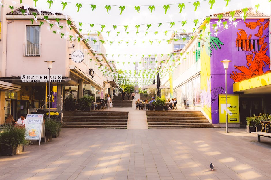 A wide outdoor pedestrian area in the town centre during daytime, featuring a series of wide concrete steps leading up to a raised seating and walking space. Bright green paper lanterns and triangular flags are strung overhead, creating a festive atmosphere. On the left side, a shop with a sign reading 'Ärztehaus' and storefront windows displays various items, while outdoor seating with tables and chairs is occupied by a few customers. To the right, a vibrant purple wall decorated with orange murals and a yellow advertising panel adds colour to the scene. Several people are seated at tables, some walking or standing on the steps, and a pigeon is seen in the foreground on the pavement. Potted plants and street lamps are positioned along the edges of the public space, which appears to be part of a town centre environment suitable for casual leisure or socialising. Man With a Van Crayford offers professional removals and moving services in this area, supporting efficient furniture transport and home relocation tasks within the town centre amid the lively surroundings.
