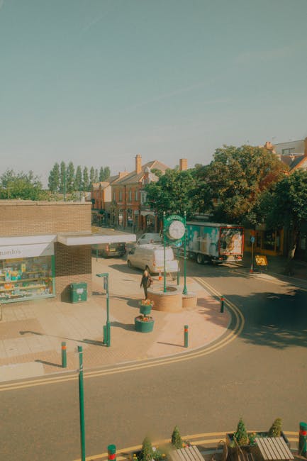 An outdoor urban scene in Crayford featuring a small, paved parking area with green bollards, planters, and a cyclist walking across the street. In the background, there are brick row houses with chimneys and pitched roofs, along with trees and a clear sky. On the left side, a small shop with a white sign displays various products, and near the shop, a green trash bin is visible. A white cargo van with an open rear door is parked on the street, and a delivery truck with blue and orange graphics is positioned further down the road. The scene is well-lit with natural daylight, and the environment appears to be in the midst of a home relocation or furniture transport process, with the area prepared for loading or unloading belongings. Man With a Van Crayford’s moving services are implied by the context of the scene involving a vehicle used for removals and home relocation tasks.