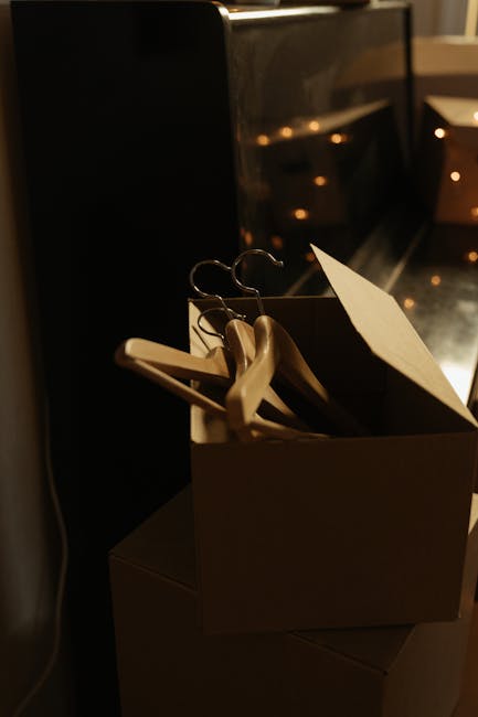 A cardboard box filled with wooden hangers and a black and white cardboard item, positioned on top of other boxes, located inside a house near a doorway or window. The hangers are hanging loosely inside the box, and some are partially visible. The scene appears to be part of a home relocation or packing process, with the boxes likely prepared for transportation or storage. The background shows a dark piece of furniture or appliance, and the lighting is warm, highlighting the packing materials and objects inside the box. This image reflects the packing and moving preparations often involved in house removals, with the focus on organized packing in readiness for a furniture transport or home relocation by Man With a Van Crayford.