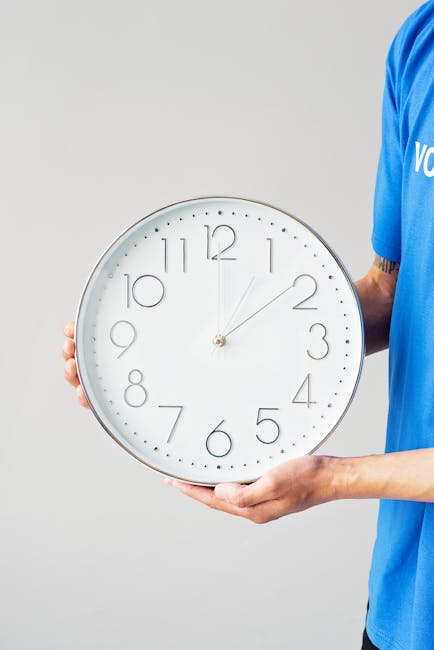 A person dressed in a blue uniform holding a large white wall clock with a silver rim and black hour markers. The clock displays a time of approximately 12:07. The person's right hand supports the clock's edge while their left hand grips the bottom, with part of their arm visible and a tattoo on the forearm. The background is plain and light grey, providing a neutral setting suitable for emphasizing the clock and the individual. This scene relates to house removals and moving services, illustrating the importance of time management and scheduling in home relocation, as provided by Man With a Van Crayford during packing and loading processes.
