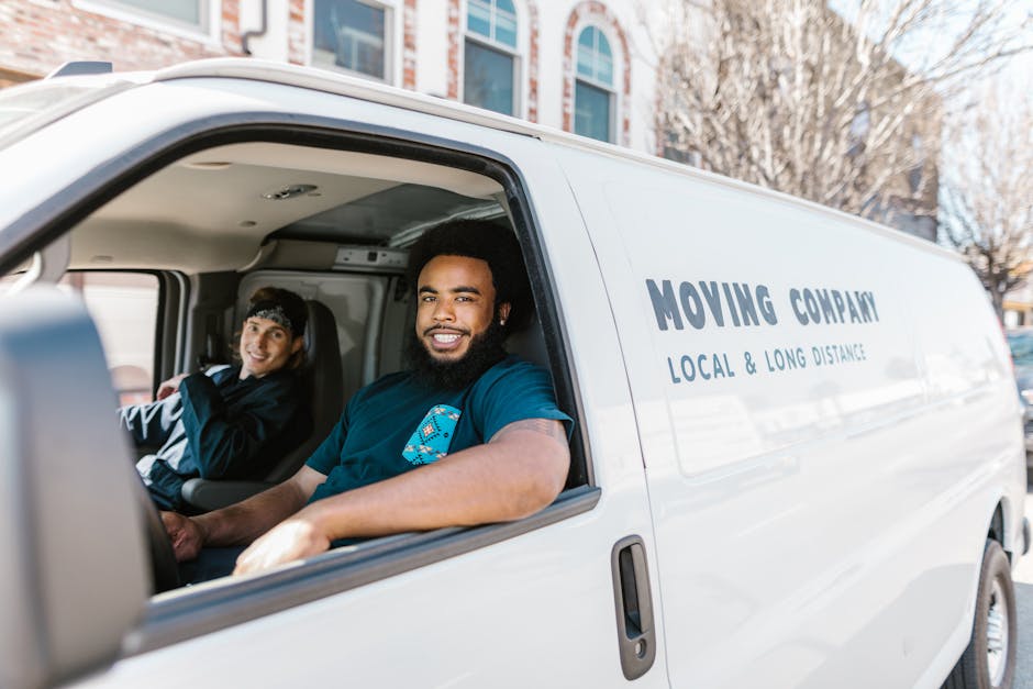 Inside the cab of a white moving van with the sliding door open, two movers are visible. The foreground features a smiling man with dark curly hair and a beard, seated in the driver's seat, wearing a 