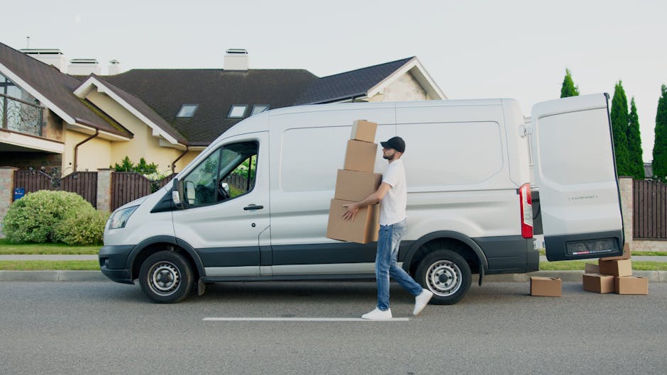 Inside the cab of a white moving van with the sliding door open, two movers are visible. The foreground features a smiling man with dark curly hair and a beard, seated in the driver's seat, wearing a 