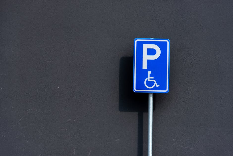 Close-up view of a designated accessible parking space on a paved surface, marked with a yellow wheelchair symbol painted on the asphalt. The parking area appears to be part of a larger parking lot wi
