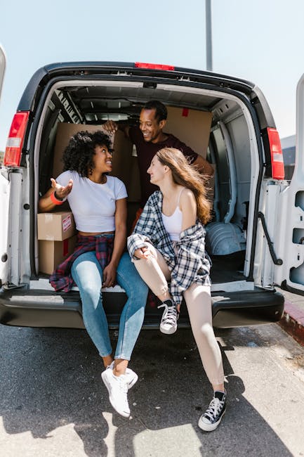 A family of three, including two young women and a man, sit inside the open rear compartment of a grey moving van during daylight. The women are seated on the edge of the van's floor, with one wearing