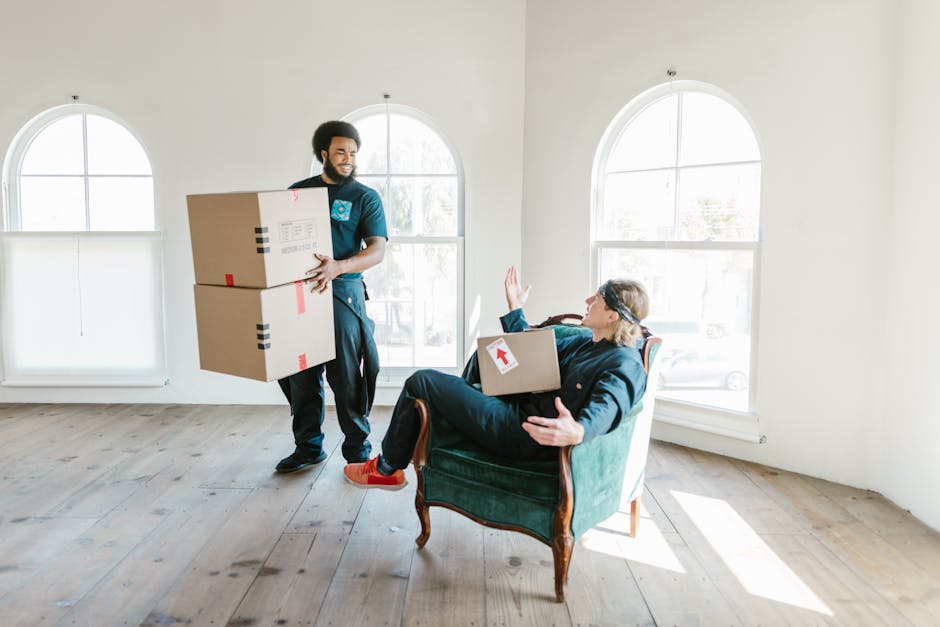 A professional mover from Man With a Van Crayford unloads two large cardboard boxes, secured with red and black tape, inside a well-lit room with white walls and wooden flooring. The mover, dressed in a dark uniform, stands near a seated individual on an elegant vintage armchair, who is holding another box with a red arrow sticker on it and raising one hand in a friendly gesture. The room features three tall, arched windows allowing natural light to flood the space, indicating a residential home during a house relocation process. The scene captures the ongoing furniture transport and packing and moving activities, with the mover carefully bringing boxes inside to facilitate a smooth home removal for a client preparing for a move, illustrating the logistics and handling involved in professional removal services.